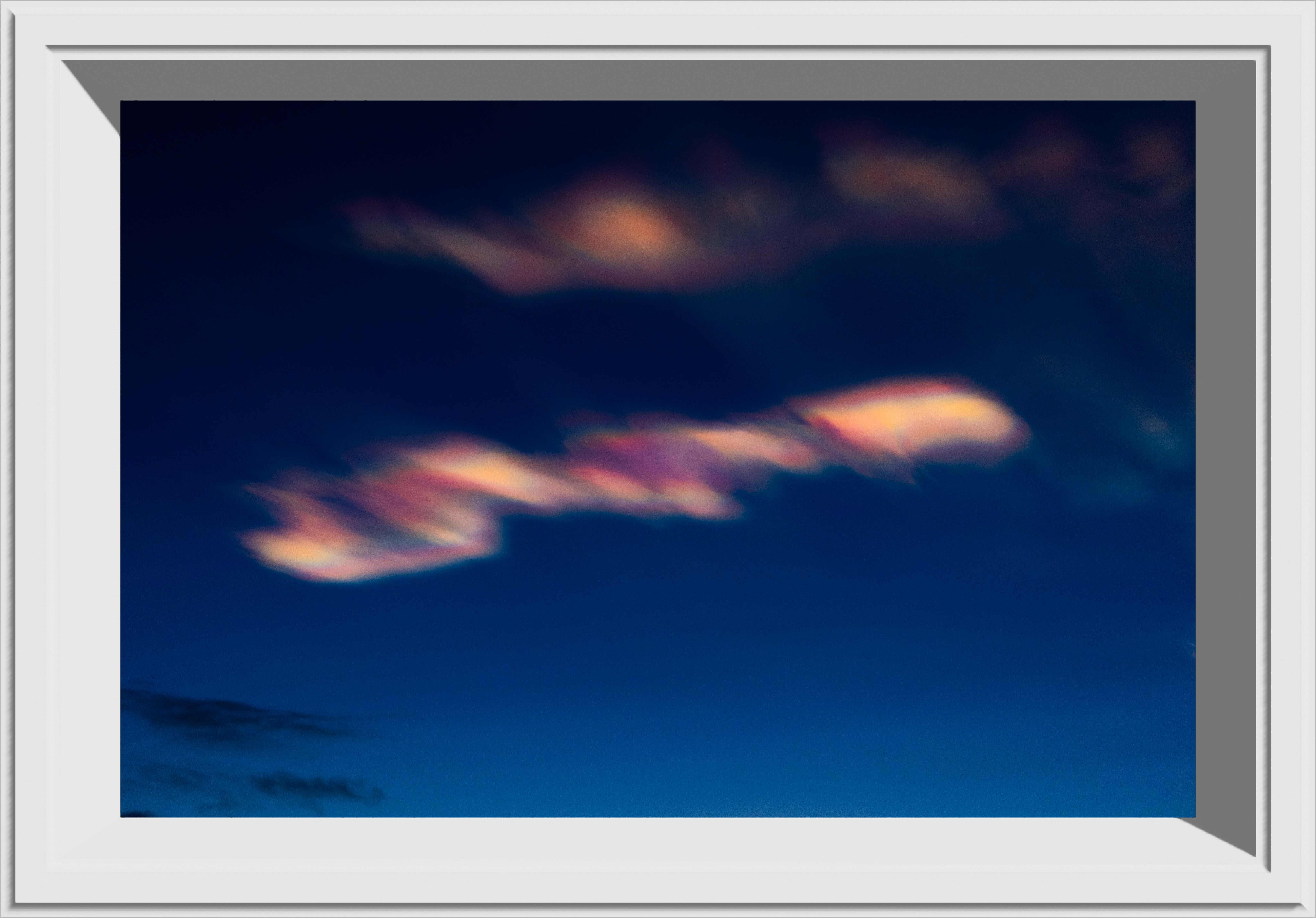 Wind scattered rainbow clouds lit by the setting sun on a dark blue background, in a white frame
