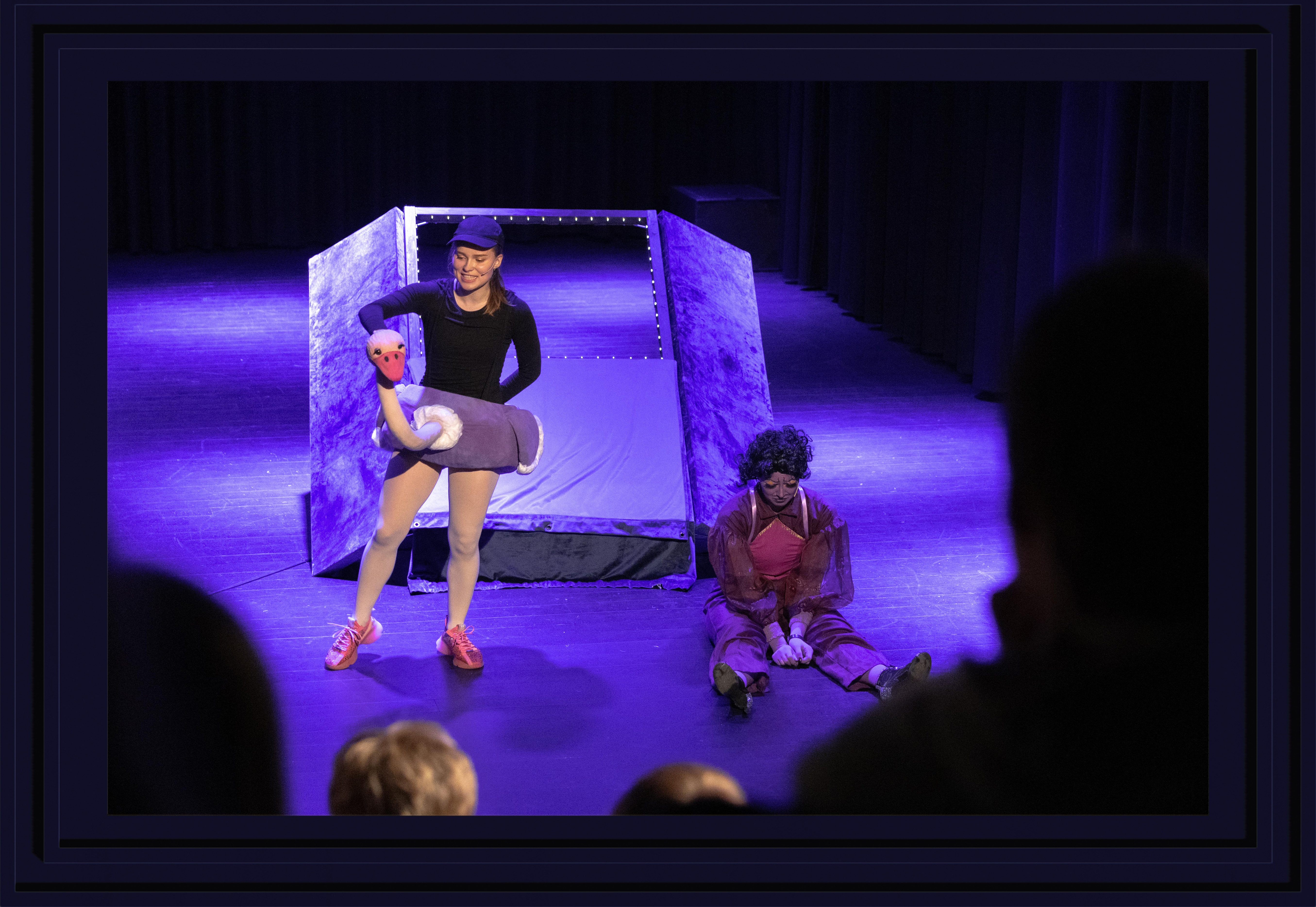 Two performers in purple light, one sitting sad in a corner the other standing in an ostrich costume trying to cheer up the other