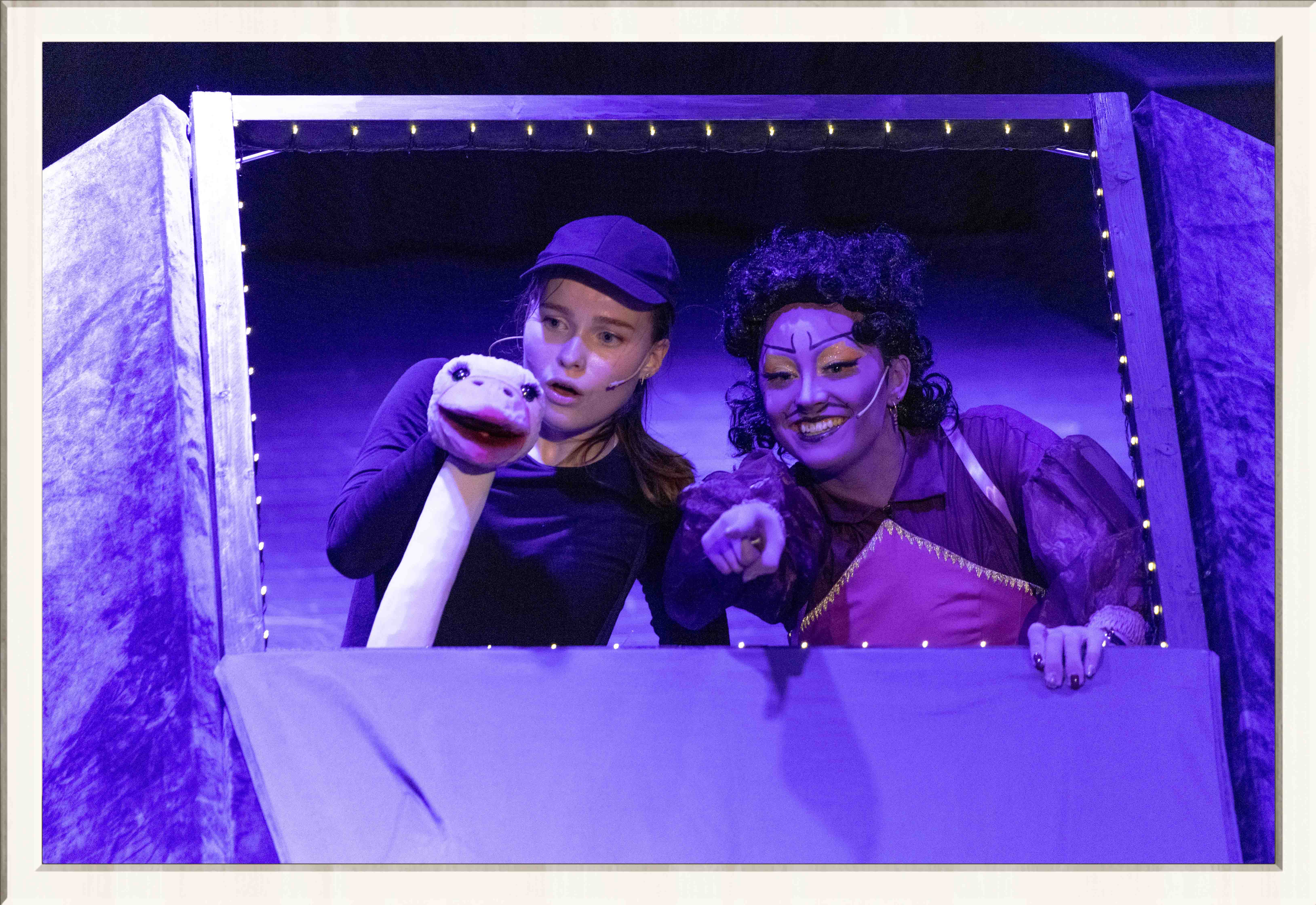 Two performers in a purple lit stage, one wearing a drag costume, the other an ostrich costume, performing a musical play for kids, framed in a thin white frame