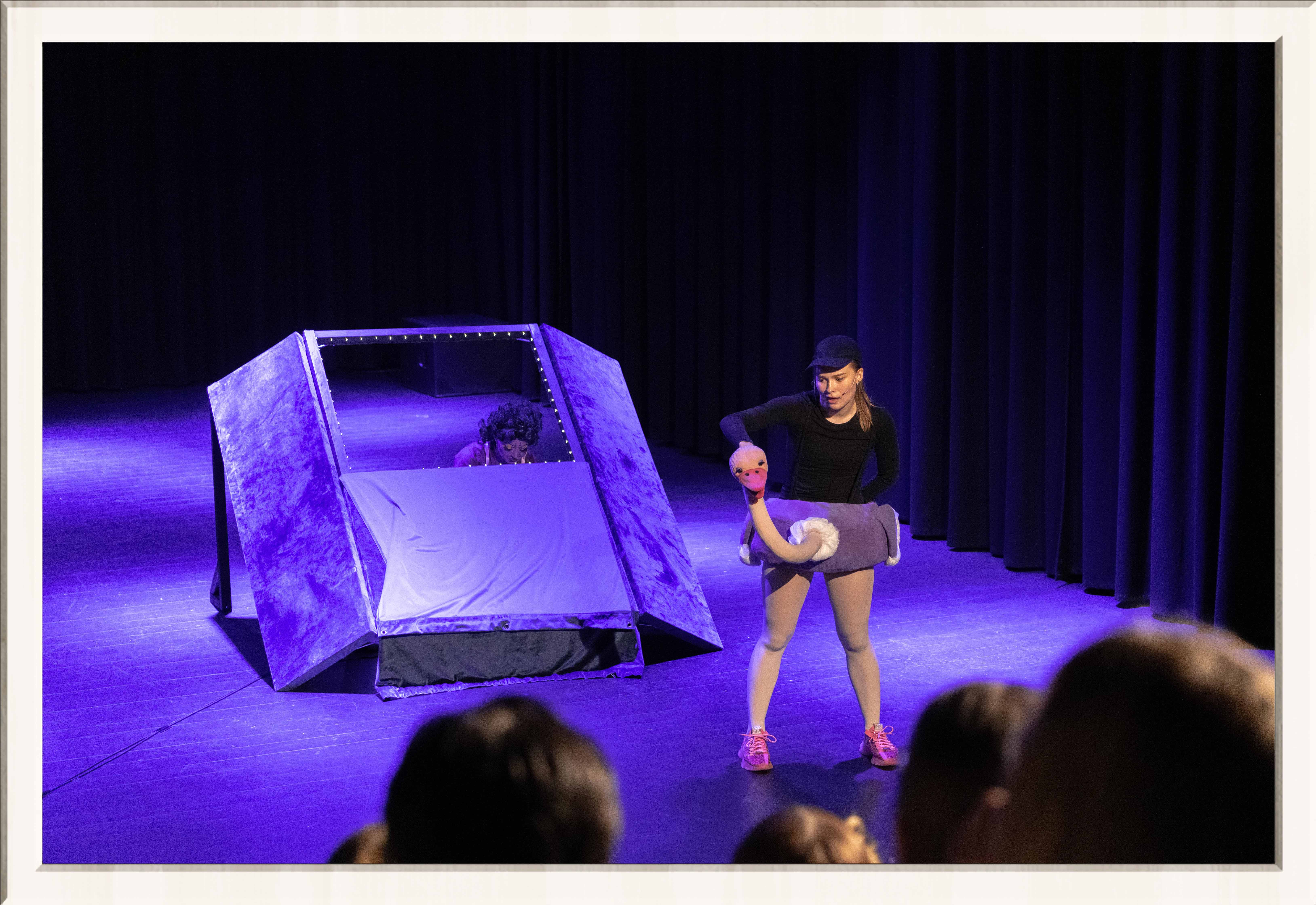 Two performers in a purple lit stage, one wearing a drag costume, the other an ostrich costume, performing a musical play for kids, framed in a thin white frame